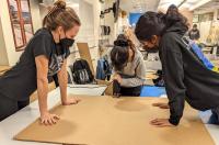 three students cutting a piece of thick cardboard in a machine shop